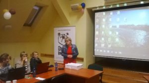 Zdjęcie przedstawia osoby słuchające szkoleniowca kobietę. W&nbsp;tle ekran projektora.