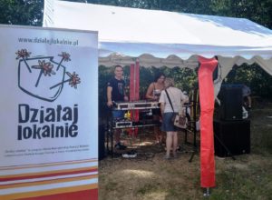 foto2 Zdjęcie przedstawia namiot, pod którym znajduje się sprzęt audio. Pod namiotem stoją trzy osoby, a obok rozłożony jest roll-up z logiem "Działaj Lokalnie"
