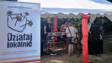 Zdjęcie przedstawia namiot, pod którym znajduje się sprzęt audio. Pod namiotem stoją trzy osoby, a obok rozłożony jest roll-up z logiem "Działaj Lokalnie"