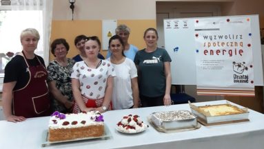 Zdjęcie przedstawia grupę osób stojącą przy stole, na którym leżą upieczone ciasta i torty.