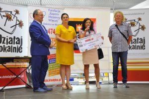 Zdjęcie przedstawia cztery odświętnie ubrane osoby. Jedna z&nbsp;nich trzyma czek na&nbsp;grant oraz&nbsp;przemawia do&nbsp;mikrofonu.