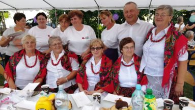 Zdjęcie przedstawia członkinie Koła Gospodyń Wiejskich wraz z innymi uczestnikami wydarzenia, pozujących do zdjęcia przy zastawionym stole.