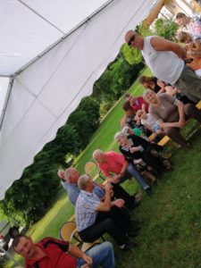 Zdjęcie przedstawia osoby siedzące na&nbsp;widowni pod&nbsp;piknikowym namiotem.