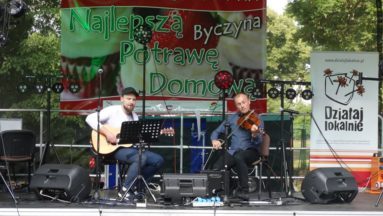 Zdjęcie przedstawia dwóch muzyków - gitarzystę i skrzypka - podczas koncertu na plenerowej scenie. W tle widać baner z logiem programu "Działaj Lokalnie" oraz baner konkursu kulinarnego "Najlepsza Potrawa Domowa".