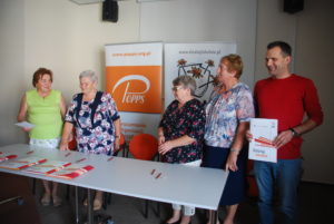 Zdjęcie przedstawia pięć os&oacute;b stojących na&nbsp;tle baner&oacute;w POPPS i&nbsp;programu Działaj Lokalnie. Jedna z&nbsp;os&oacute;b trzyma folder z&nbsp;logiem i&nbsp;nazwą programu. Na&nbsp;pierwszym planie znajduje się stolik, na&nbsp;kt&oacute;rym leżą ułożone foldery.