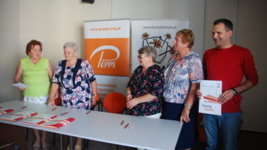 Zdjęcie przedstawia pięć osób stojących na tle banerów POPPS i programu Działaj Lokalnie. Jedna z osób trzyma folder z logiem i nazwą programu. Na pierwszym planie znajduje się stolik, na którym leżą ułożone foldery.