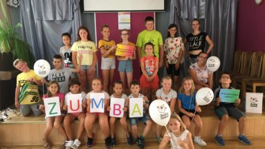 Zdjęcie przedstawia grupę dzieci na sali gimnastycznej, pozujących do zdjęcia. Dzieci w pierwszym rzędzie trzymają kartki tworzące napis ZUMBA, a kilkoro innych dzieci trzyma balony z logiem "Działaj Lokalnie".