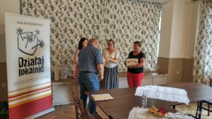Zdjęcie przedstawia cztery osoby stojące w&nbsp;pomieszczeniu. Jednej z&nbsp;nich wręczany jest symboliczny czek oraz&nbsp;torba
