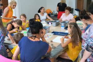 Na zdjęciu widać duży st&oacute;ł, na&nbsp;kt&oacute;rym rozłożone są farby, kartki papieru oraz&nbsp;kamienie. Dookoła stołu siedzą dzieci oraz&nbsp;dorośli, przygotowujący się do&nbsp;warsztat&oacute;w plastycznych.