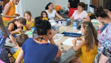 Na zdjęciu widać duży stół, na którym rozłożone są farby, kartki papieru oraz kamienie. Dookoła stołu siedzą dzieci oraz dorośli, przygotowujący się do warsztatów plastycznych.