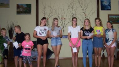 Zdjęcie przedstawia grupę dzieci i młodzieży stojącą na scenie. W tle widać wiszące na ścianie obrazy i zdjęcia.