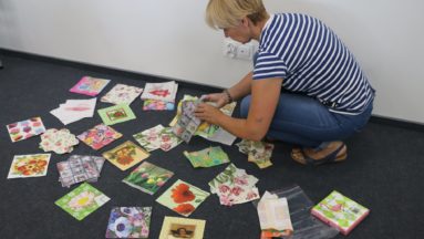 Zdjęcie przedstawia instruktorkę zajęć kucającą wśród rozłożonych na podłodze kartek z dziełami powstałymi podczas warsztatów decoupage.