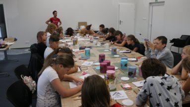 Zdjęcie przedstawia grupę dzieci siedzącą przy dużym stole podczas warsztatów decoupage.