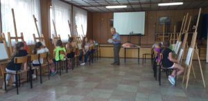 Zdjęcie przedstawia widok wnętrza. Po&nbsp;obu stronach sali dzieci siedzą w&nbsp;rzędzie przy&nbsp;sztalugach, malując; pomiędzy dwoma rzędami sztalug stoi instruktor zajęć.