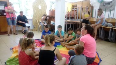 Na zdjęciu widzimy grupę dzieci wraz z dwoma instruktorkami zajęć, siedzącymi w kole na kolorowym kocu w pomieszczeniu.