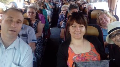 Zdjęcie przedstawia wnętrze autokaru, w którym siedzą uśmiechnięci uczestnicy wycieczki.