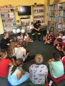 Zdjęcie przedstawia grupę dzieci siedzących w&nbsp;k&oacute;łku na&nbsp;podłodze w&nbsp;bibliotece. Obok&nbsp;na krześle siedzi mężczyzna w&nbsp;strażackim stroju, pokazujący dzieciom książkę i&nbsp;opowiadający o&nbsp;niej.