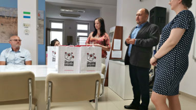 Zdjęcie przedstawia pięć osób stojących przy stole we wnętrzu. Na stole stoją torby z logiem programu "Działaj Lokanie".