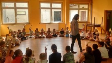 Zdjęcie przedstawia grupę dzieci siedzącą w kole w sali gimnastycznej. W środku stoi osoba prowadząca zajęcia.
