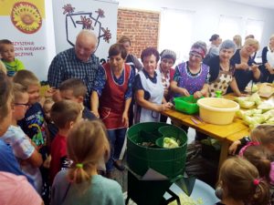 Na zdjęciu widać grupę ludzi w&nbsp;pomieszczeniu - starsze osoby pokazują młodszym, jak szatkować kapustę.