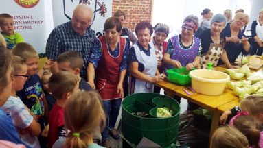 Na zdjęciu widać grupę ludzi w pomieszczeniu - starsze osoby pokazują młodszym, jak szatkować kapustę.