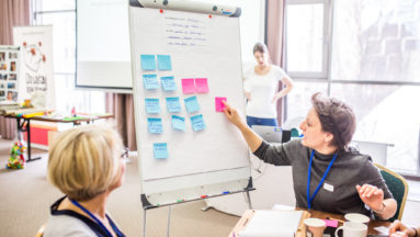 Zdjęcie przedstawia dwie osoby dyskutujące podczas warsztatów. Jedna z nich nakleja karteczki post-it na flipcharta.