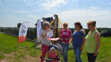 Zdjęcie przedstawia grupę dorosłych i dziecko na polu, nieopodal baneru "Działaj Lokalnie"