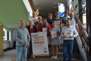 Zdjęcie przedstawia grupę os&oacute;b w&nbsp;pomieszczeniu, pozujących do&nbsp;zdjęcia, trzymających plakat z&nbsp;napisem "Wyzwolisz społeczną energię"