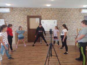 Na zdjęciu widzimy grupę dzieci i&nbsp;instruktorkę podczas zajęć sportowych. Na&nbsp;środku sali stoi aparat na&nbsp;statywie, za&nbsp;pomocą kt&oacute;rego nagrywane są zajęcia.