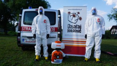 Dwie osoby w kombinezonach i maseczkach, stojące na tle baneru z logiem "Działaj Lokalnie"