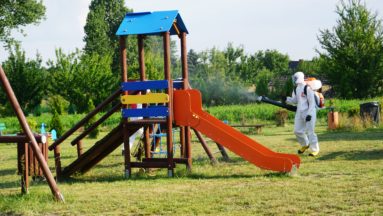 Dwóch wolontariuszy w kombinezonach dezynfekuje plac zabaw.