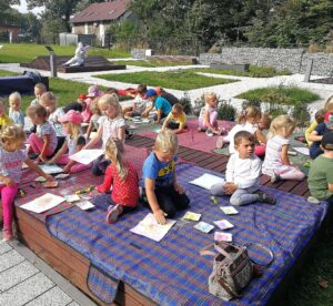 dzieci na&nbsp;matach piknikowych tworzą prace plastyczne, w&nbsp;tle Kicak odpoczywa na&nbsp;ławce