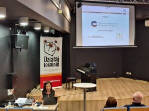 Koordynator programu przestawiający prezentację w&nbsp;ramach programu Działaj Lokalnie, widoczne logotypy programu oraz&nbsp;roll up Działaj Lokalnie.