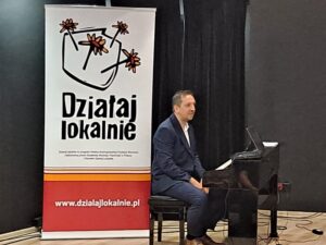 Muzyk siedzący za&nbsp;pianinem w&nbsp;tle roll up Działaj Lokalnie