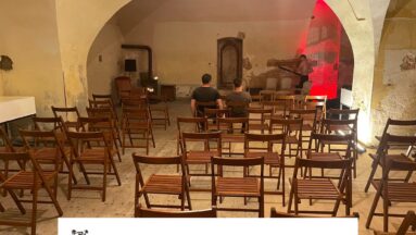 Na zdjęciu widać dużą ilość drewnianych krzeseł. W tle widać mężczyznę z odkurzaczem. Dwóch mężczyzn siedzi na krzesłach.